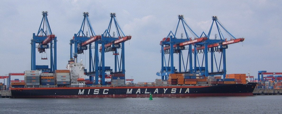 A container ship berthed at a terminal, with stacks of containers and gantry cranes alongside